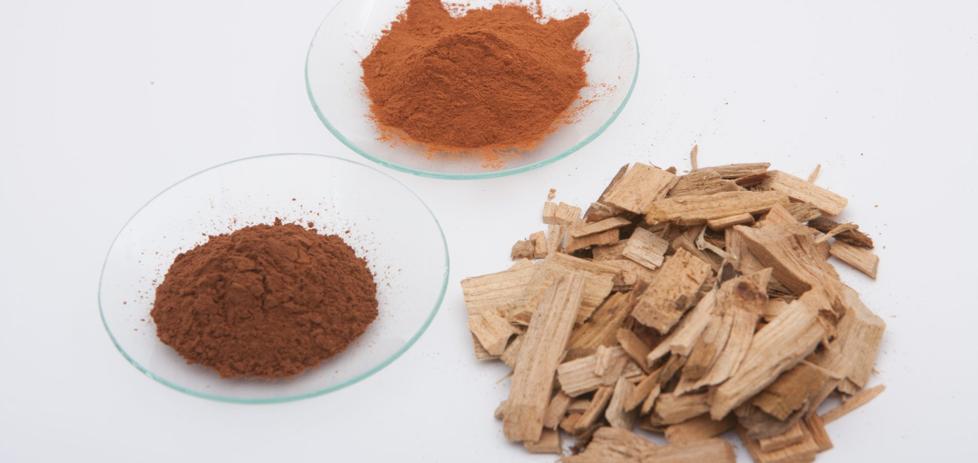 Due piastre di vetro con polvere marrone-rossastra accanto a un mucchio di scaglie di legno chiaro su sfondo bianco.
